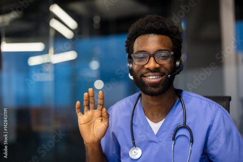 Obraz na płótnie A smiling healthcare professional wearing a headset waves hello, seemingly ready to assist. The doctor sports glasses and a stethoscope. w salonie