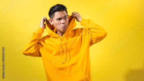 sian man in bright yellow hoodie making a funny expression while pulling hoodie collar with both hands, standing against vibrant yellow background, great for humor, casual fashion, or reaction visuals