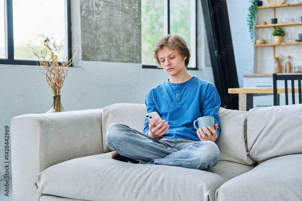 Relaxed teenager guy sitting at home on the couch with smartphone and cup of tea