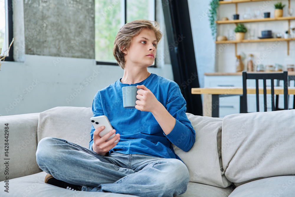 Relaxed teenager guy sitting at home on the couch with smartphone and cup of tea