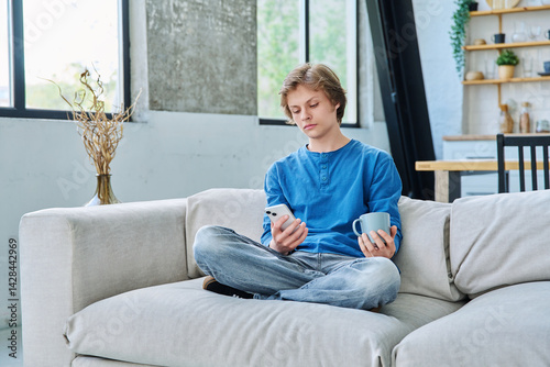 Obraz na płótnie Relaxed teenager guy sitting at home on the couch with smartphone and cup of tea w salonie