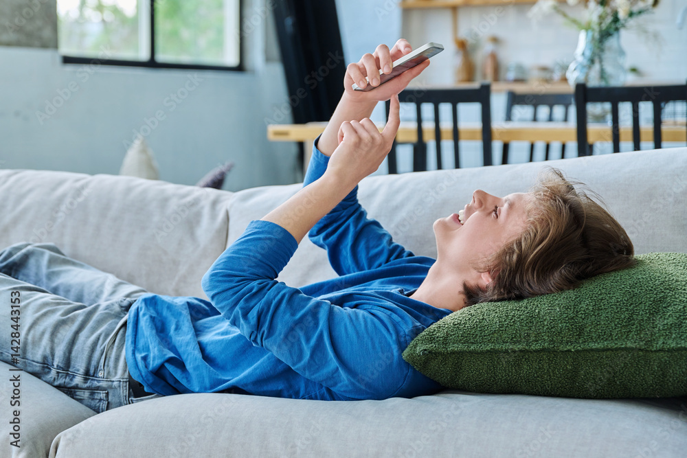 Happy joyful handsome teenager guy using smartphone lying on the couch at home