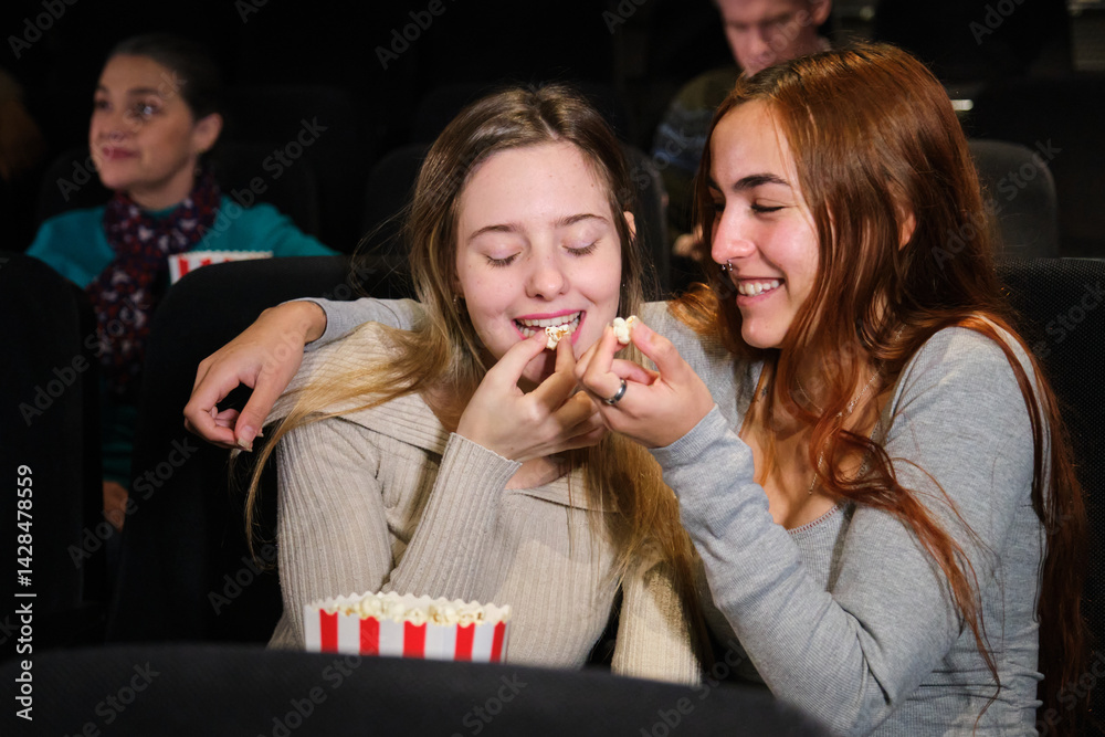 Two friends are watching a movie and eating popcorn, having a good time together at the cinema