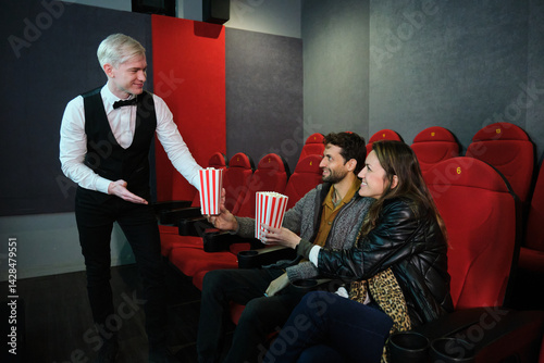 Obraz na płótnie Elegant waiter serving popcorn to a young couple sitting in the red seats of a movie theater w salonie