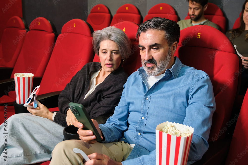 Senior couple using phone while watching movie at cinema, with popcorn and 3d glasses