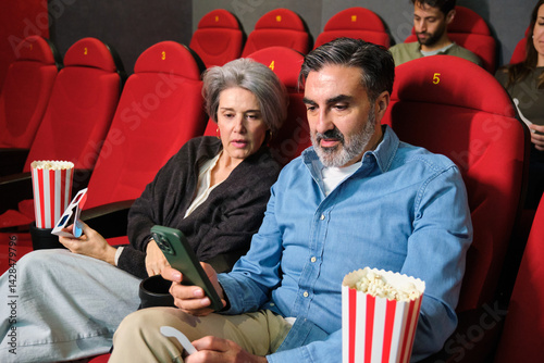 Obraz na płótnie Senior couple using phone while watching movie at cinema, with popcorn and 3d glasses w salonie