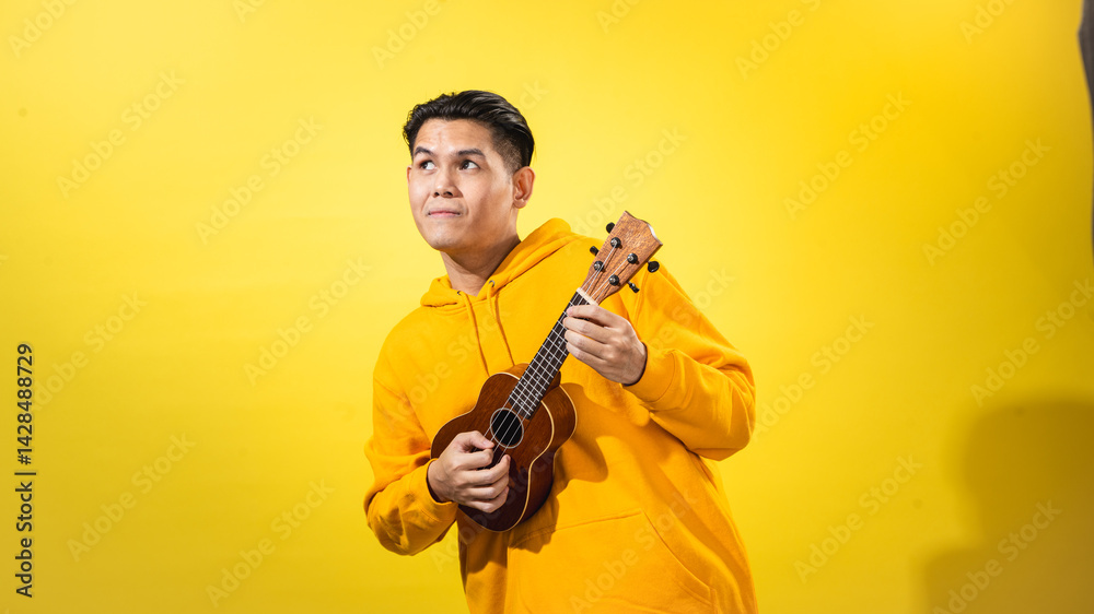 Asian man in yellow hoodie enthusiastically playing a small ukulele against a vivid yellow background. Expressive musical portrait with happy mood and creative, artistic personality