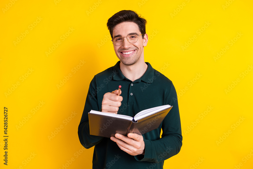 Handsome young man with glasses holding a notebook and pen, smiling confidently against a vibrant yellow background Handsome young man with glasses holding a notebook and pen, smiling confidently against a vibrant yellow background