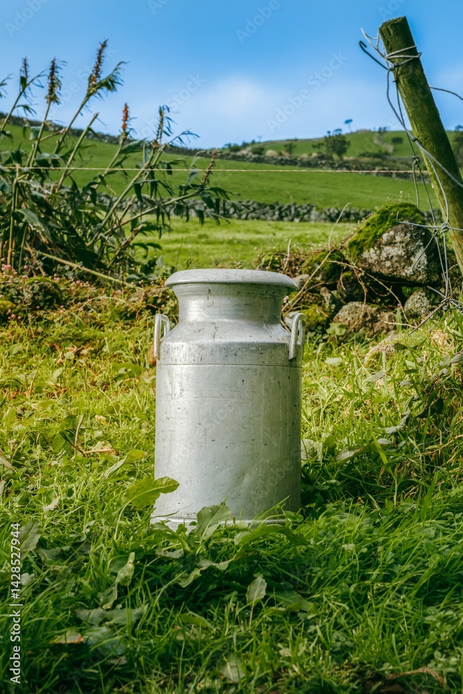 A aluminum milk churn rests in a field of lush green grass, Vintage Milk Churn in a Lush Green Pasture Azores. Rustic Aluminum Milk Can Standing Amidst the Vibrant Countryside of Ilha de Sao Jorge