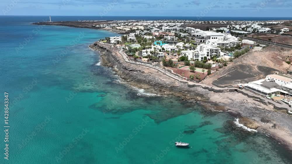 Panoramaflug entlang der Küste von Playa Blanca, Lanzarote: türkisfarbenes Meer, schwarze Felsen, Hotels, Palmen und vulkanische Landschaften aus der Vogelperspektive.
