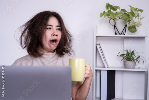 Tired Asian woman sitting at her office desk, yawning and holding a cup of coffee, feeling sleepy and low energy in the morning due to a sleepless night, hangover, or trouble falling asleep