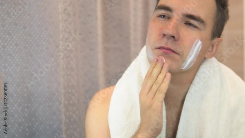 A young guy with a white jersey applies shaving foam to his face and looks in the mirror. Bathroom with daylight. Male skin care.