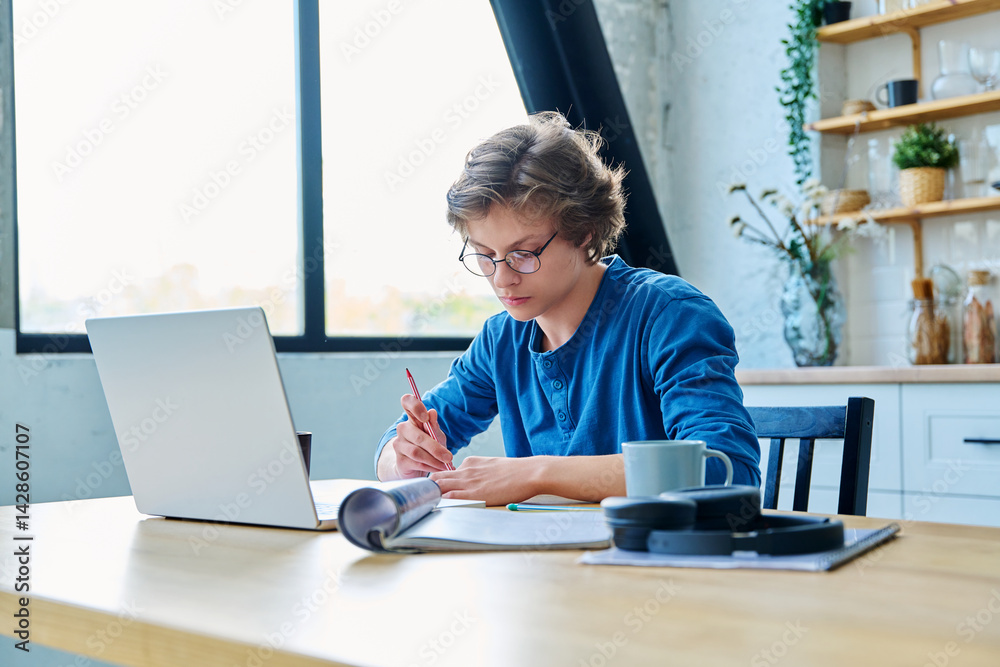Teenage guy student sitting at the table with laptop, textbooks in home interior