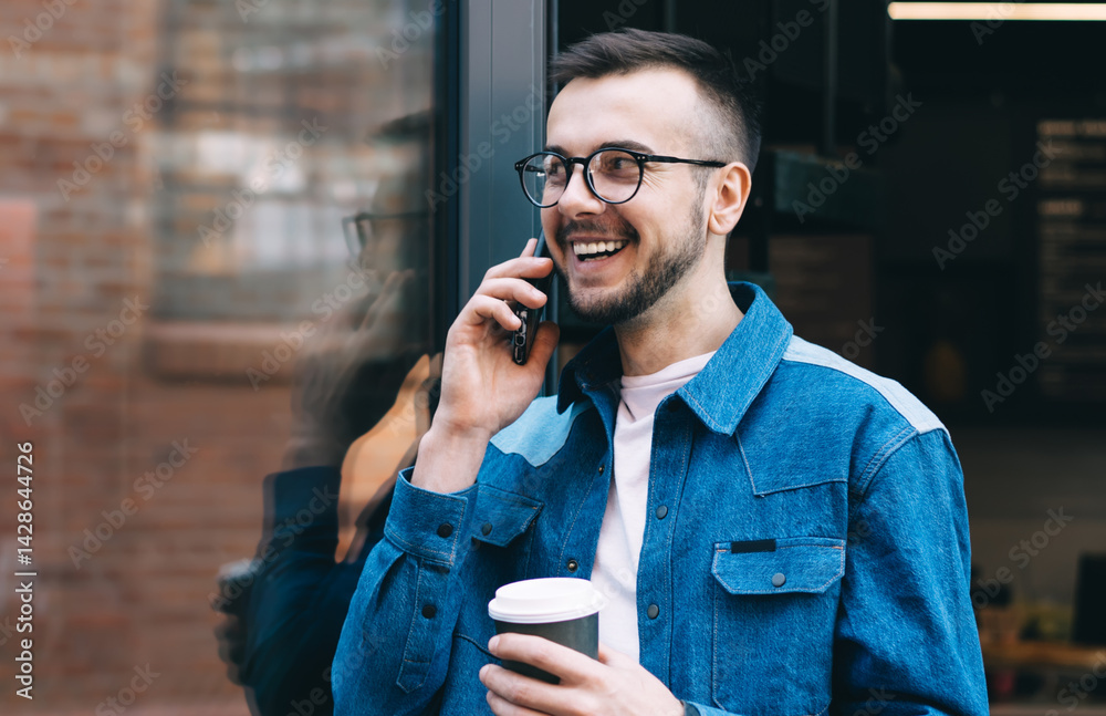 Cheerful male student talking on smartphone while standing with coffee near glass door of coffee shop