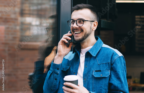 Obraz na płótnie Cheerful male student talking on smartphone while standing with coffee near glass door of coffee shop w salonie