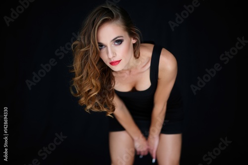 Fashionable sexy woman posing in black mini dress, looking at the camera with delicate smile. Studio shot. Glamour makeup. Wavy long hair. Fashionable sexy woman posing in black mini dress, 