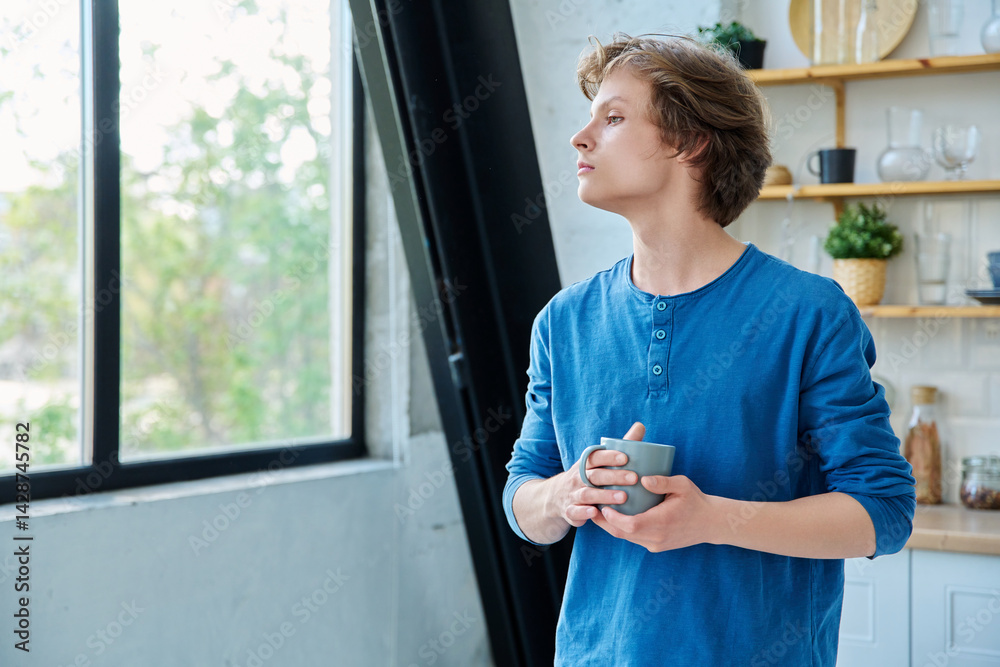 Teenage serious guy with cup in hands in home interior, looking out window
