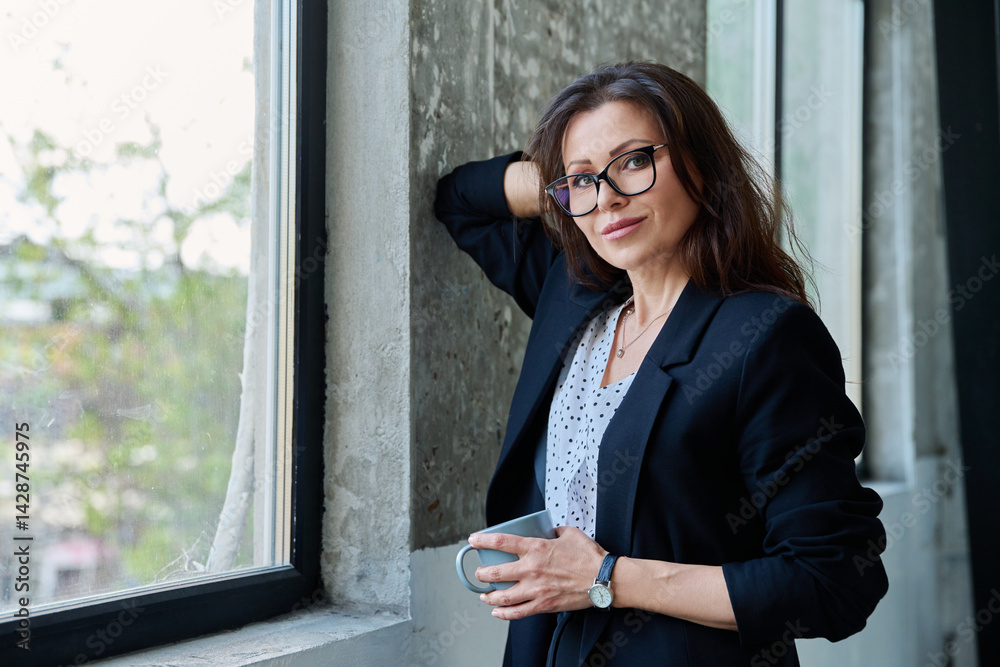 Portrait of middle aged beautiful businesswoman looking at camera, standing near window