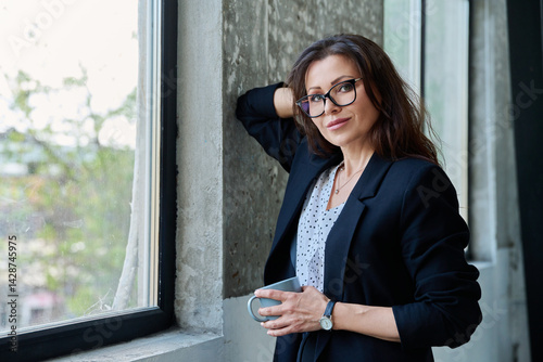 Obraz na płótnie Portrait of middle aged beautiful businesswoman looking at camera, standing near window w salonie