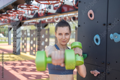 A woman in a sports top holds dumbbells outside.The woman in the photo expresses confidence and strength. Her posture and gaze demonstrate determination and willingness to train.
