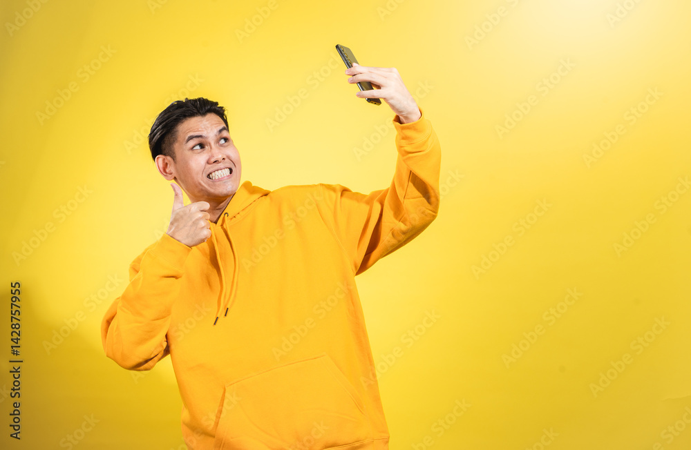 Playful young Asian man in yellow hoodie taking selfie while giving thumbs-up and making a funny face, set against vibrant yellow background. Studio shot representing joyful digital expression