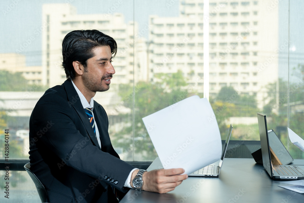 South Asian businessman holding business paper while talking during meeting with Caucasian businesswoman seated across office desk with laptops showing corporate engagement and communication exchange South Asian businessman holding business paper while talking during meeting with Caucasian businesswoman seated across office desk with laptops showing corporate engagement and communication exchange