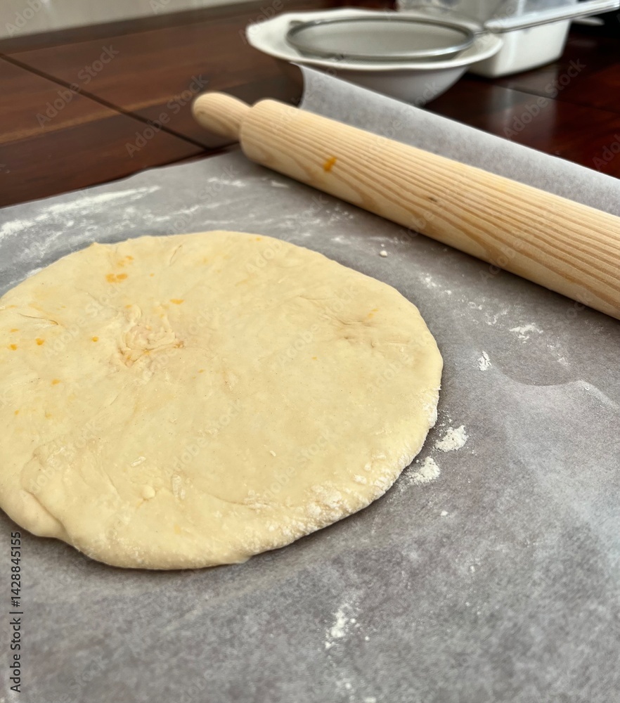 A round piece of dough with a wooden dough roller on a wooden board with baking parchment paper sprinkled with flour. Sieve flour, muffin, cake, roll out dough. Sift. Cooking process A round piece of dough with a wooden dough roller on a wooden board with baking parchment paper sprinkled with flour. Sieve flour, muffin, cake, roll out dough. Sift. Cooking process