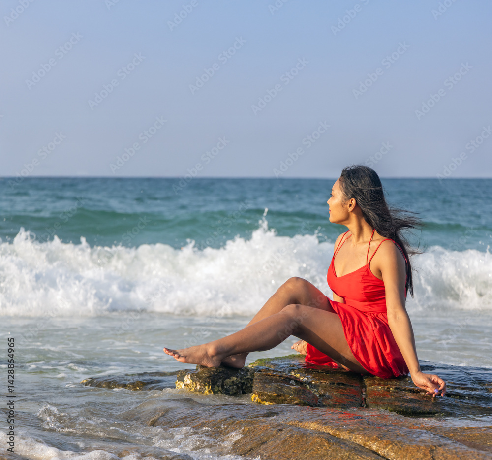 Woman in red dress sits on a rock on the coast and watches the sea