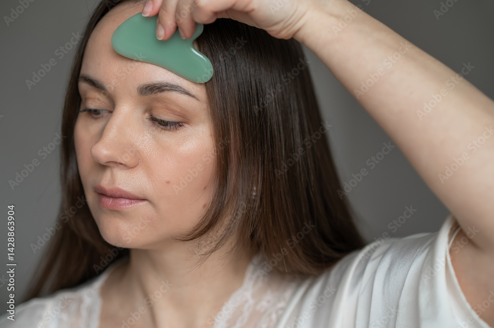 Naklejka premium Young woman using a green gua sha scraper for facial massage, improving skin circulation and relaxation. Home skincare and self-care routine for beauty and wellness.