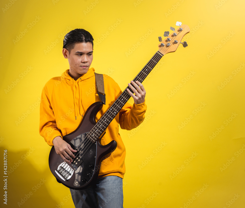 Asian man in a yellow hoodie playing an electric bass guitar against a bright yellow background. Captured mid-performance with a focused expression and casual modern style