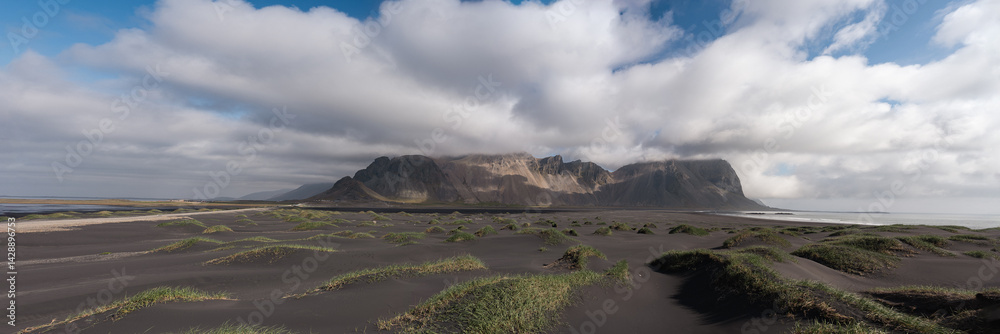 Wide-angle shot of Vestrahorn mountains emerging from mist with soft light illuminating black sand dunes and sparse vegetation on the Icelandic coast.