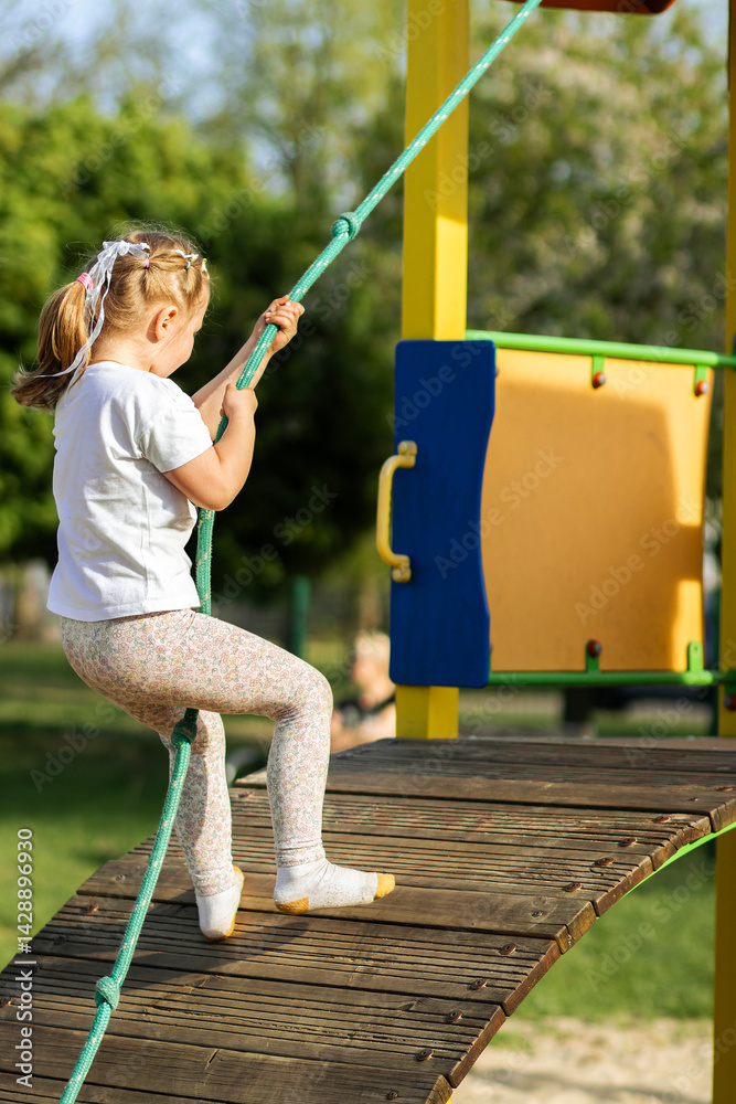 Little girl in white shirt and floral leggings climbs a wooden playground bridge using a green rope, with colorful equipment and greenery in the sunny background.