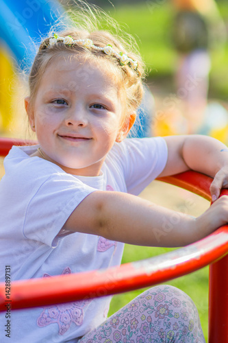 Obraz na płótnie Happy blonde girl in a white T-shirt with a floral headband smiling joyfully on a vibrant, colorful playground. The background is full of playful red, yellow, green, and blue elements. w salonie