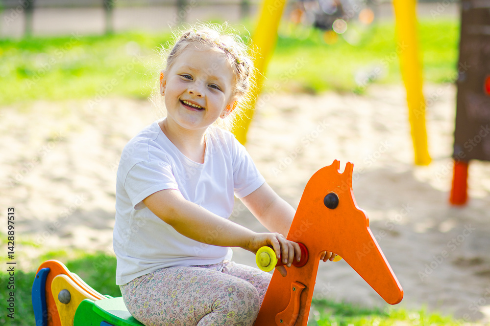 Smiling little girl in white shirt and patterned leggings rides an orange spring rocker shaped like a horse on a colorful playground with grass and sand in the background.
