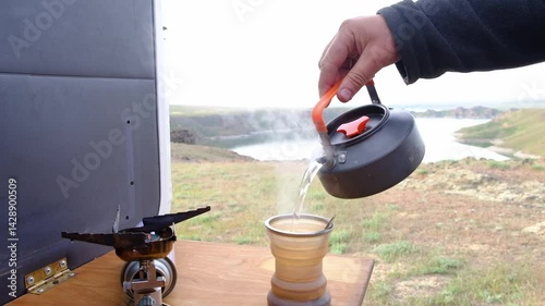 Morning tea is prepared in a kettle in a camper car on a gas stove with a beautiful view of the lake and rocks.