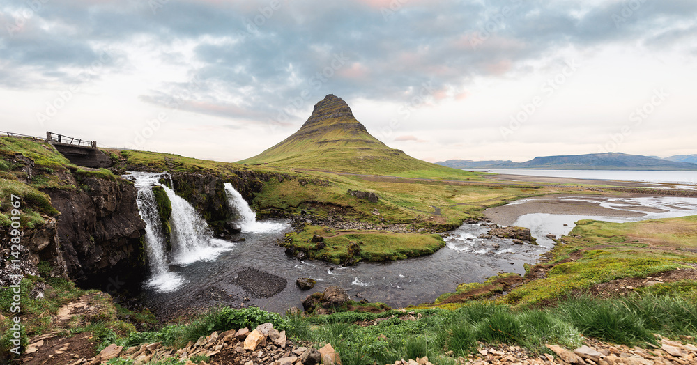 Panoramic view of Kirkjufell mountain with a waterfall in the foreground, surrounded by lush green fields and a river flowing toward the Icelandic coast.