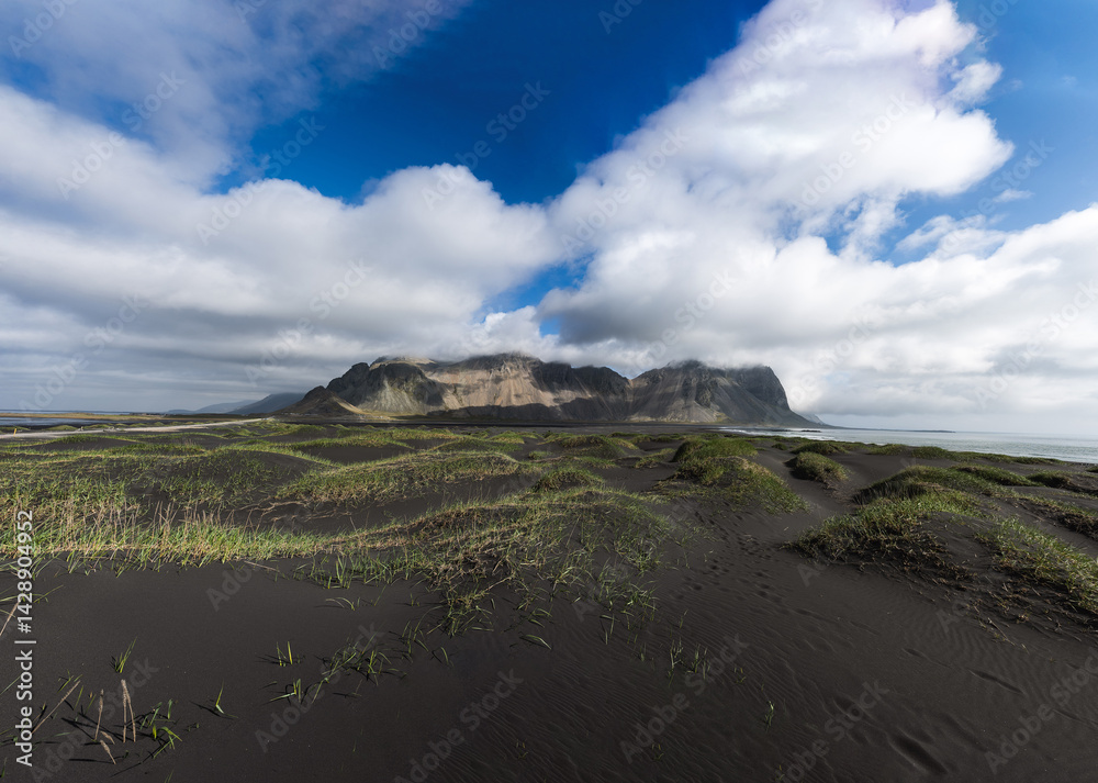 Dramatic panoramic view of Vestrahorn mountain with dark sand dunes and scattered grass tufts under a cloudy sky in Iceland.