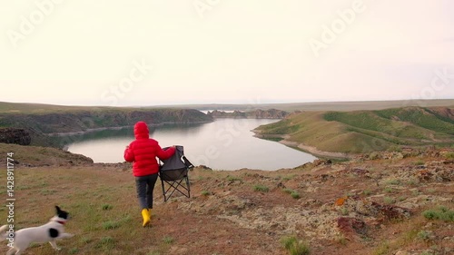 A woman or girl in a red down jacket and yellow EVA boots carries a tourist chair closer to the shore of a beautiful lake with rocks to admire the beautiful evening view, accompanied by a black and wh