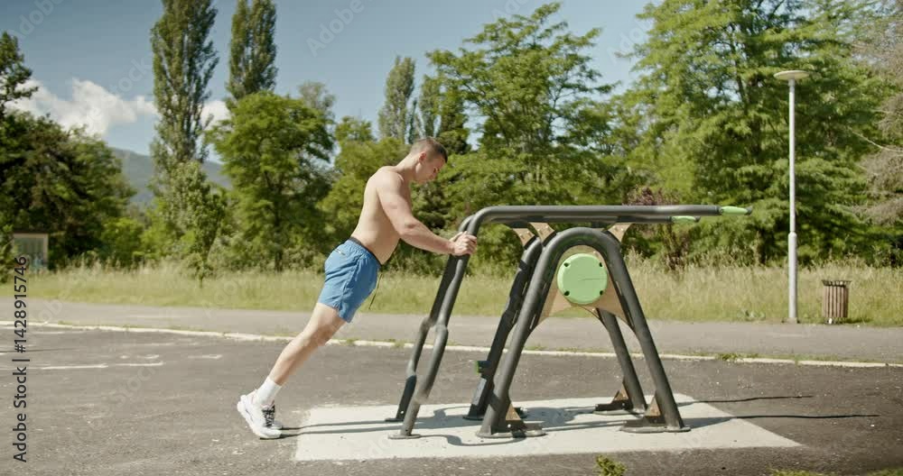 A muscular man performs incline pushups on an outdoor workout machine in a park. He completes his set and walks away.