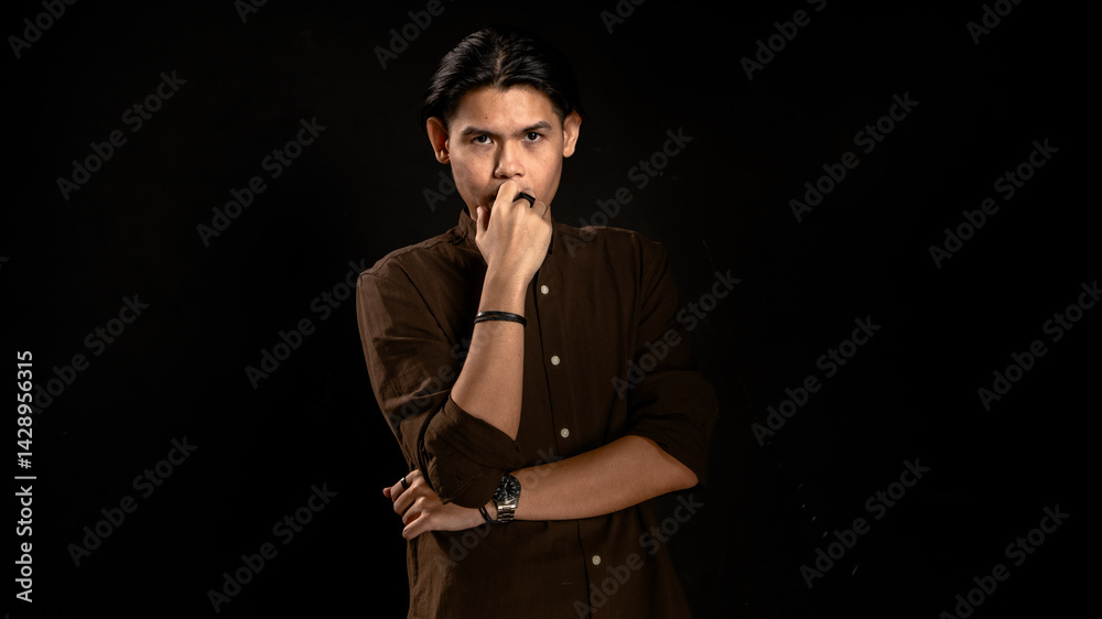 Asian man in dark brown shirt standing in studio with dark background, touching lips with thoughtful expression, perfect for concept visuals related to thinking, planning, or personal reflection