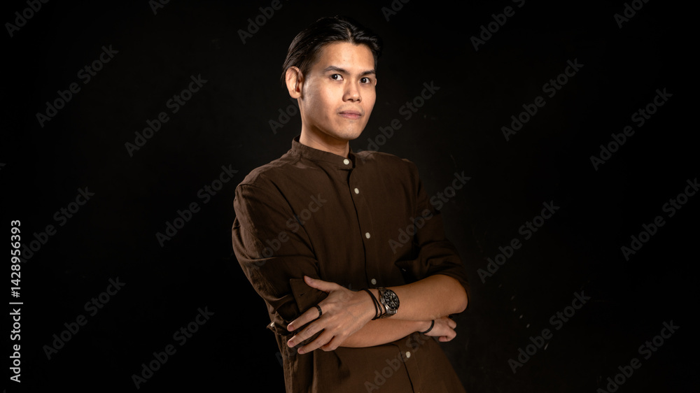 Asian man wearing dark brown button-up shirt standing confidently with arms crossed in front of dark studio background, suitable for lifestyle, leadership, or minimalist personal branding visuals