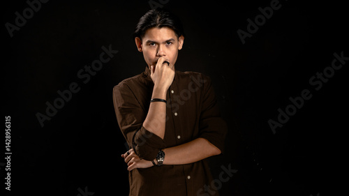 Obraz na płótnie Asian man in dark brown shirt standing in studio with dark background, touching lips with thoughtful expression, perfect for concept visuals related to thinking, planning, or personal reflection w salonie