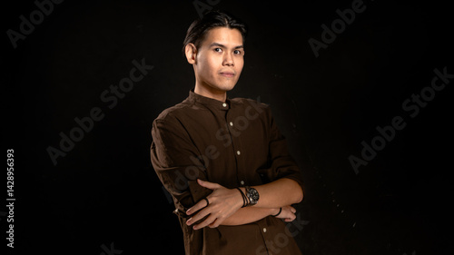 Obraz na płótnie Asian man wearing dark brown button-up shirt standing confidently with arms crossed in front of dark studio background, suitable for lifestyle, leadership, or minimalist personal branding visuals w salonie