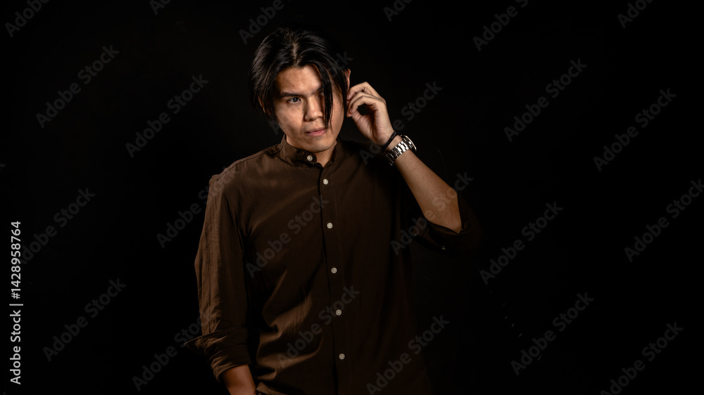 Asian man in dark brown button-up shirt standing in studio with hand touching ear and intense gaze, posing confidently against black background, ideal for fashion, branding, or edgy lifestyle content