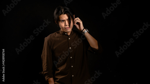 Obraz na płótnie Asian man in dark brown button-up shirt standing in studio with hand touching ear and intense gaze, posing confidently against black background, ideal for fashion, branding, or edgy lifestyle content w salonie