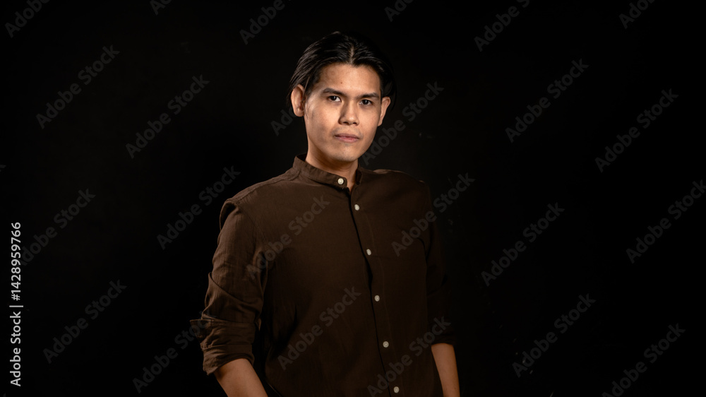 Asian man wearing dark brown button-up shirt posing with a relaxed and confident look in studio against dark background, suitable for personal branding, fashion, or minimalist creative content
