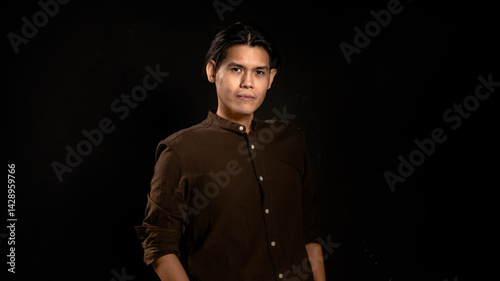 Obraz na płótnie Asian man wearing dark brown button-up shirt posing with a relaxed and confident look in studio against dark background, suitable for personal branding, fashion, or minimalist creative content w salonie