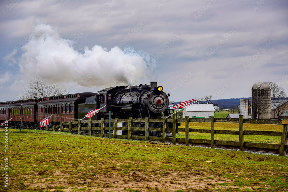 Obraz premium Historic steam locomotive pulls vintage passenger cars along tracks while puffs of steam rise against a cloudy sky. The setting features a green field and wooden fence nearby.
