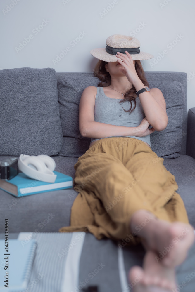 Obraz premium Young woman lying on a cozy sofa, hiding her face with a stylish straw hat, surrounded by books, a camera, and headphones, savoring moments of relaxation and tranquility at home
