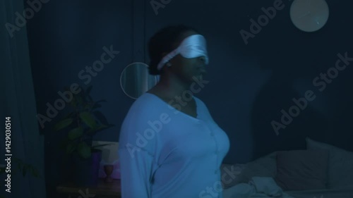 Slow motion shot of young African American woman in satin eye mask and pajamas standing in dark bedroom, moving from side to side while sleepwalking at night